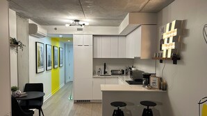 Een moderne keuken met witte kasten, een ingebouwde oven en een magnetron. Er staan twee zwarte barkrukken bij een klein aanrecht met een spoelbak.