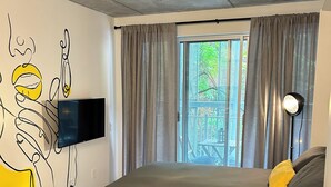 Een slaapkamer met een bed, een stoel, een aan de muur bevestigde televisie en een raam met gordijnen.