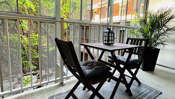 Een balkon met een tafel en stoelen, een bloempot en uitzicht op groen en gebouwen.