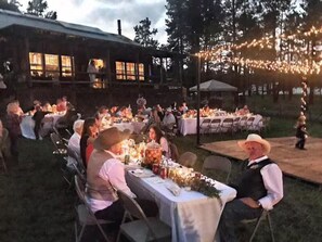 An outdoor dining area with tables set for guests, surrounded by a rustic building and string lights.