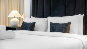 Una cama bien tendida con sábanas blancas y dos almohadas oscuras, una lámpara de noche blanca y un cabecero oscuro.