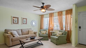 A living room with a beige sofa, two armchairs, a coffee table, a ceiling fan, and a window with curtains. - Lemon Drop Beach Cottage (Navarre)