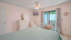 A bedroom with a bed, a ceiling fan, a dresser, a chair, a television, and a window with curtains. - Lemon Drop Beach Cottage (Navarre)