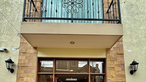 Une entrée d’immeuble avec une porte en verre, un balcon doté d’une rambarde en métal et des murs dont le motif rappelle la pierre.
