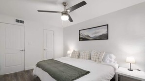 Ein Schlafzimmer mit Bett, Deckenventilator, Nachttisch mit Lampe und einem gerahmten Bild an der Wand.