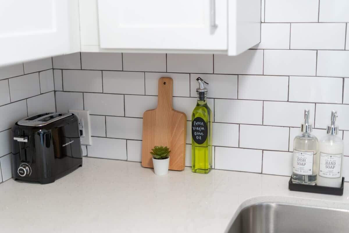 A kitchen with white tiled backsplash, a toaster, a wooden cutting board, a small potted plant, and bottles of dish and hand soap.