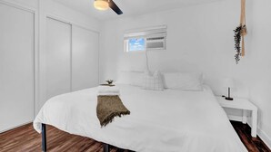 A bedroom with a bed, a ceiling fan, a small window with an air conditioner, and a bedside table with a lamp. - King Bed | Close to Chase Field (Phoenix)