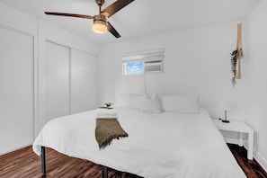 Une chambre à coucher comprenant un lit, un ventilateur de plafond, une petite fenêtre avec un climatiseur et une table de chevet avec une lampe.