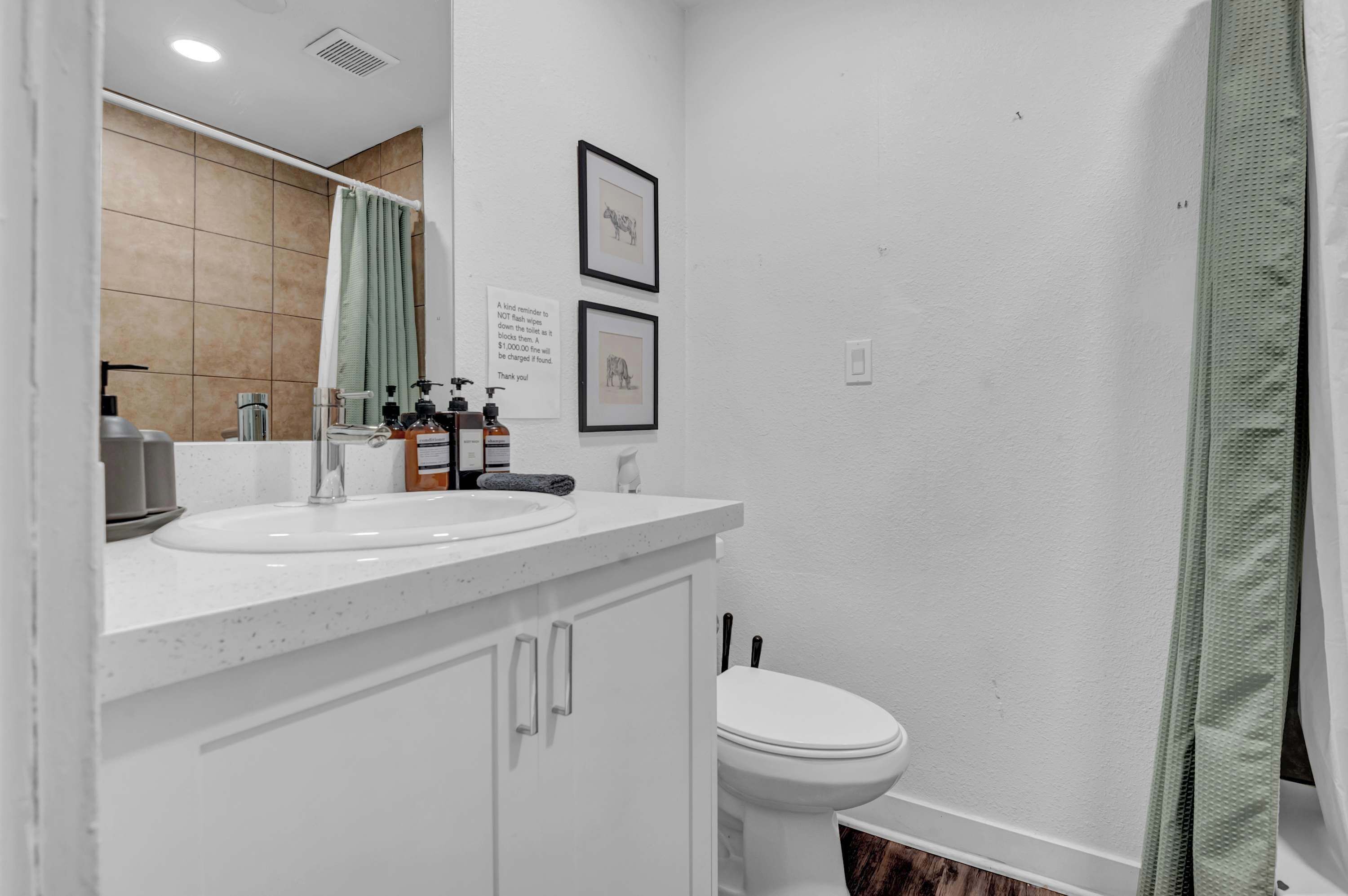 A modern bathroom with a white sink, mirror, and toilet. There are framed pictures on the wall and a green shower curtain.