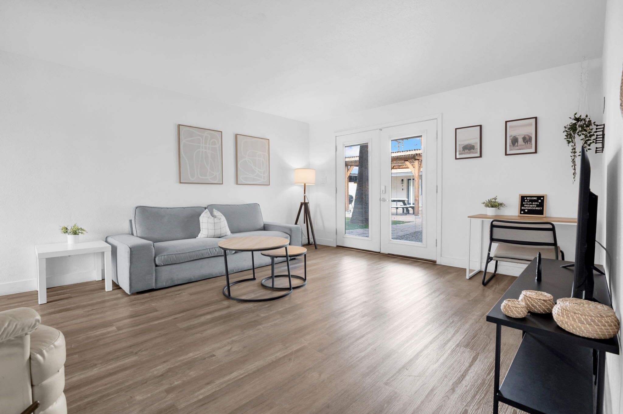 Un salon moderne avec un canapé gris, une table basse ronde et une petite table d’appoint. On y trouve une lampe, un téléviseur et un bureau avec une chaise. Le sol est en bois et les murs sont blancs.