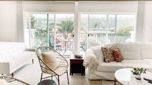 A bright, spacious room with a white sofa, a wicker chair, a small table, and a balcony with a view of greenery and buildings. - Hotel California" La Costa Resort Life (Carlsbad)
