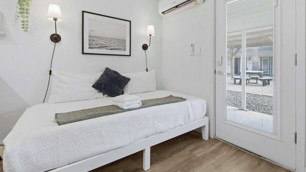 Een net opgemaakt bed met een wit hoofdeinde, een donkere kussen en een witte deken. Aan weerszijden van een lijst hangen twee wandlampen. Boven het bed is een airconditioningunit bevestigd aan de muur.