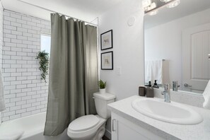 Un baño moderno con pared de azulejos blancos, cortina de ducha, inodoro, lavabo con espejo y planta en maceta.