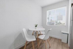 Una habitación pequeña y luminosa con una mesa blanca y cuatro sillas blancas, una ventana con persianas y una puerta blanca.