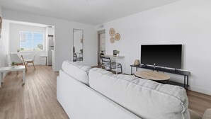 Ett modernt vardagsrum med en stor vit soffa, en matplats med ett vitt bord och stolar, en tv som är monterad på väggen och ett fönster med persienner.