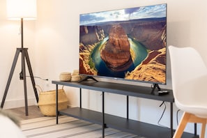 A modern living room with a flat-screen TV mounted on a black stand, a floor lamp, and a white chair. - 1 King Bed in Midtown | Near Golf (Phoenix)