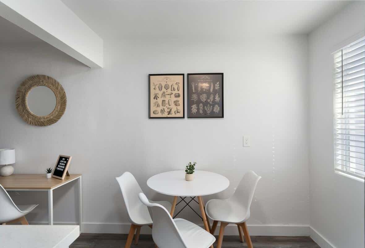 A minimalist dining area with a round table, four white chairs, a small potted plant, and two framed botanical prints on the wall.