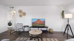 A modern living room with a flat-screen TV, a round coffee table, a sofa, a chair, a lamp, and a wall-mounted plant. - Midtown 2 King| Near Roosevelt Row (Phoenix)