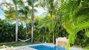 Ein Swimmingpool mit Liegestühlen und einer Hängematte, umgeben von Palmen und viel Grün.