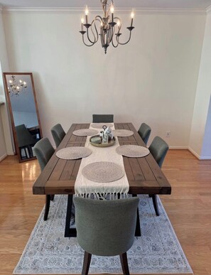 Comedor con mesa de madera, sillas grises, un mantel tejido, una lámpara de araña y un espejo.