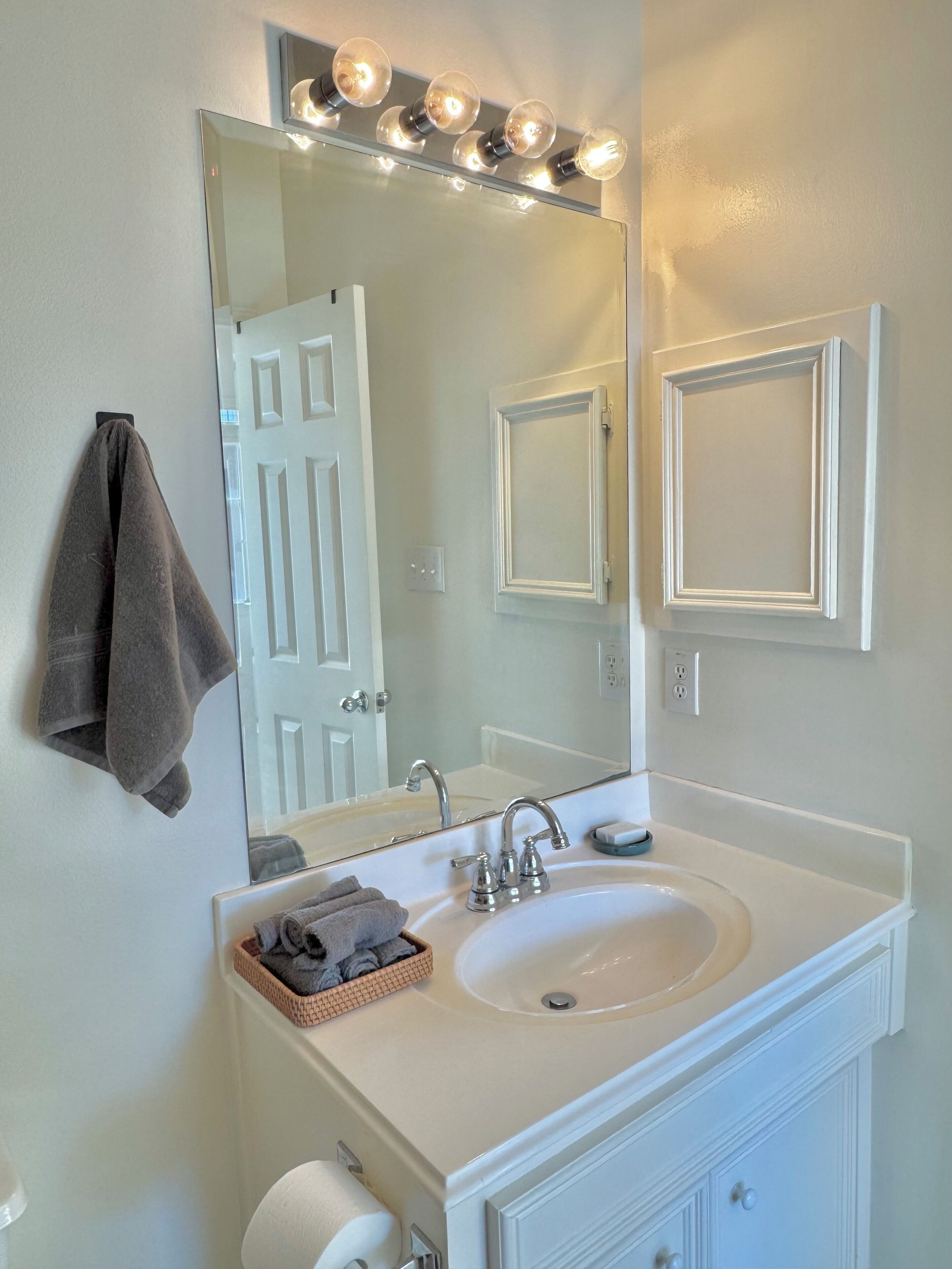 A bathroom with a white sink, a mirror with lights, a towel rack, and a door.
