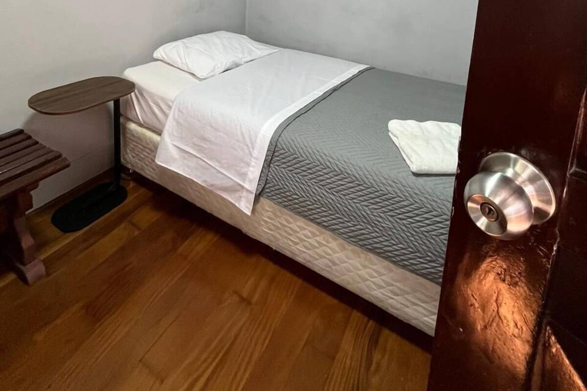 Egy kis, egyágyas szoba, fa padlóval, egy kis asztallal és egy ezüst színű kilincses ajtóval.