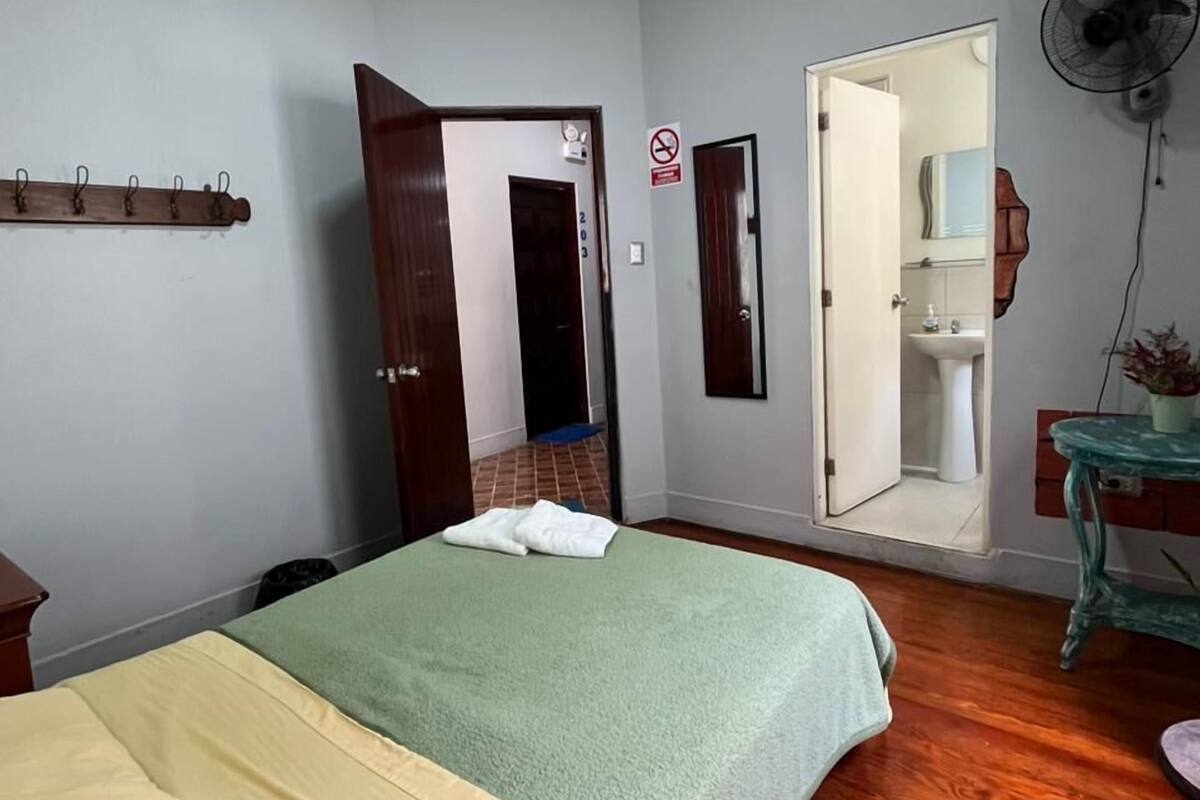 Egy hálószoba, amelyben van egy ágy, parketta, egy a falra szerelt ventilátor, és egy ajtó, amely a fürdőszobába vezet.