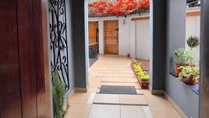A narrow corridor with a tiled floor, a wooden door at the end, and a view of a red flowered garden through an archway. - Haku Hostel Lima (Lima)