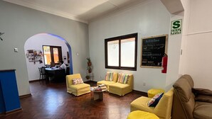 A living room with a wooden herringbone floor, yellow armchairs, a brown sofa, and a small coffee table. There is a chalkboard on the wall listing activities and a fire extinguisher mounted on the wall. - Haku Hostel Lima (Lima)