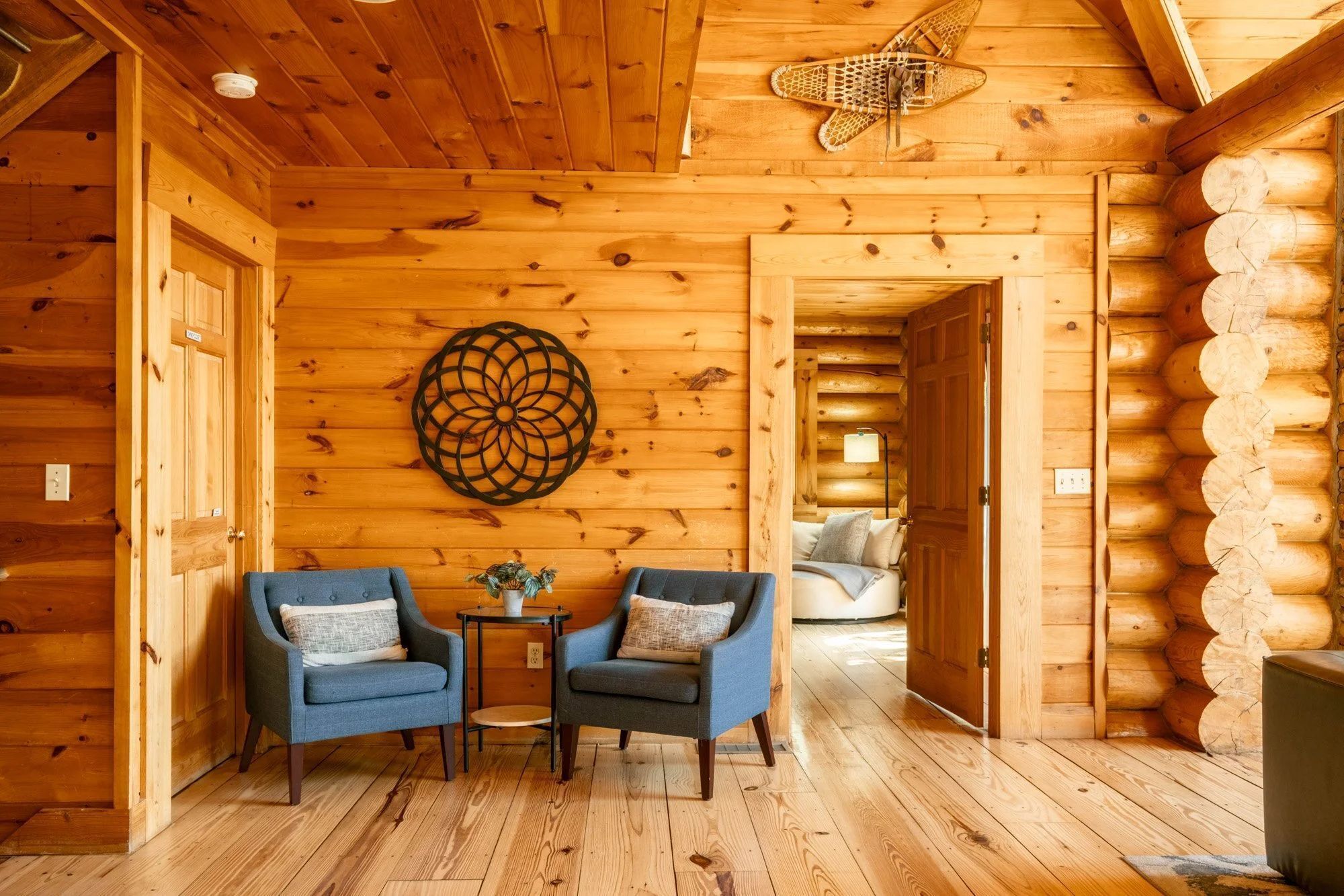 A log cabin interior with two blue armchairs, a small table, a decorative wall piece, and a visible door leading to another room.