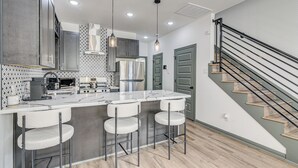 A modern kitchen with a central island, bar stools, stainless steel appliances, and a staircase with a metal railing. - Sleek, Convenient New-Build 8 Mi to Dtwn Houston! (Houston)