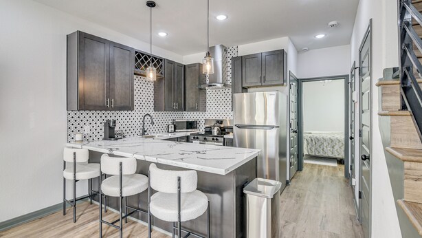 A modern kitchen with a central island, bar stools, stainless steel appliances, and a staircase leading to another level. - Sleek, Convenient New-Build 8 Mi to Dtwn Houston! (Houston)