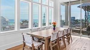 Een eethoek met een houten tafel en stoelen, grote ramen met uitzicht op de oceaan en het strand, en een glazen deur naar een terras.