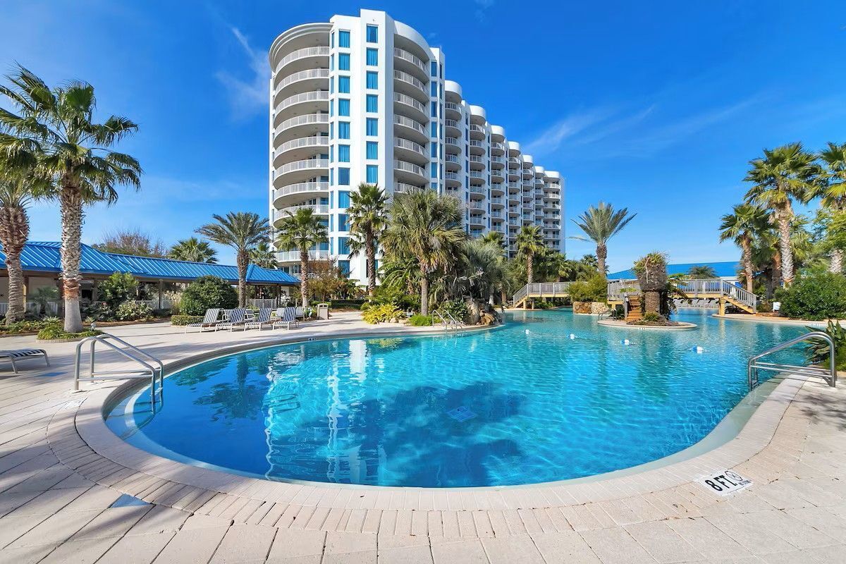A hotel with a large pool, palm trees, and a clear blue sky.
