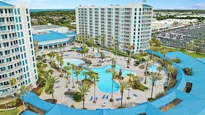 A resort with multiple high-rise buildings, a central pool area surrounded by palm trees, and a parking lot. - 2026 Updated 2BR | Largest Lagoon Size Pool (Destin)