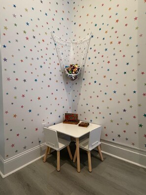 A room with a white wall featuring a star pattern, a white table with two chairs, and a hanging basket with toys.
