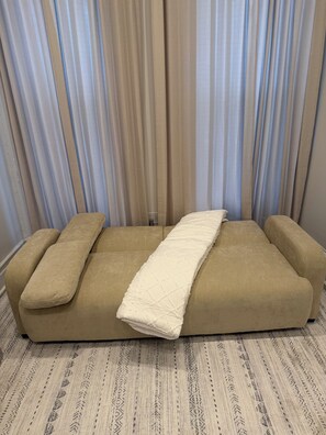 Un sofá con un cojín acolchado en una habitación con alfombra estampada y cortinas translúcidas.