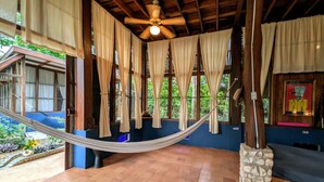 Una habitación con hamaca, vigas de madera, ventilador de techo y ventanas grandes con cortinas.