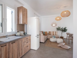 A modern kitchen with wooden cabinets, a white refrigerator, and a dining area with a sofa and table. - Pounta | 2BR Apartment w/ Shared Pool | Fiora I (24Q5+MPP Pounta, Greece)