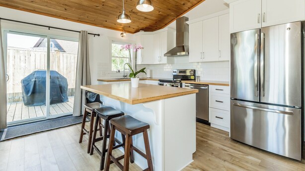 Une cuisine moderne avec des meubles blancs, un réfrigérateur en acier inoxydable, un plafond en bois et un grand îlot central doté de tabourets.