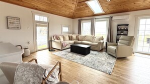 Un salon spacieux avec un plafond en bois, un canapé beige, une table basse noire et une grande fenêtre.