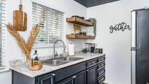 A modern kitchen with dark cabinets, a granite countertop, a stainless steel refrigerator, and a window with blinds. - The Hide-Out! A Boho Cabin AC- Wi-fi (Mi-Wuk Village)