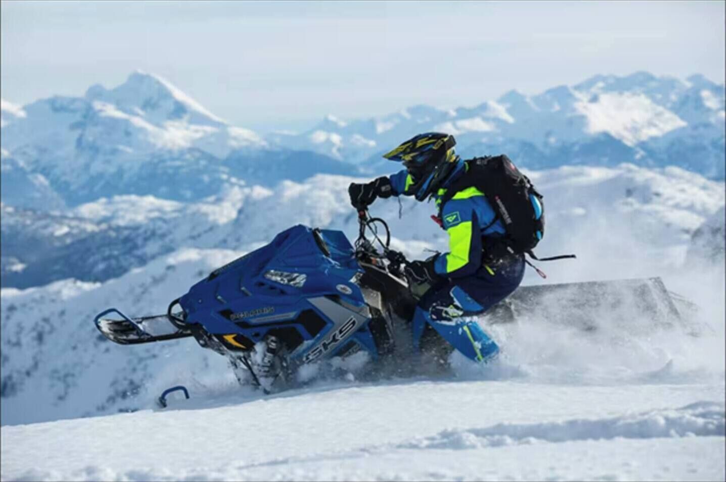 Una persona montando en moto de nieve, vestida con equipo azul y amarillo, sobre una montaña nevada.