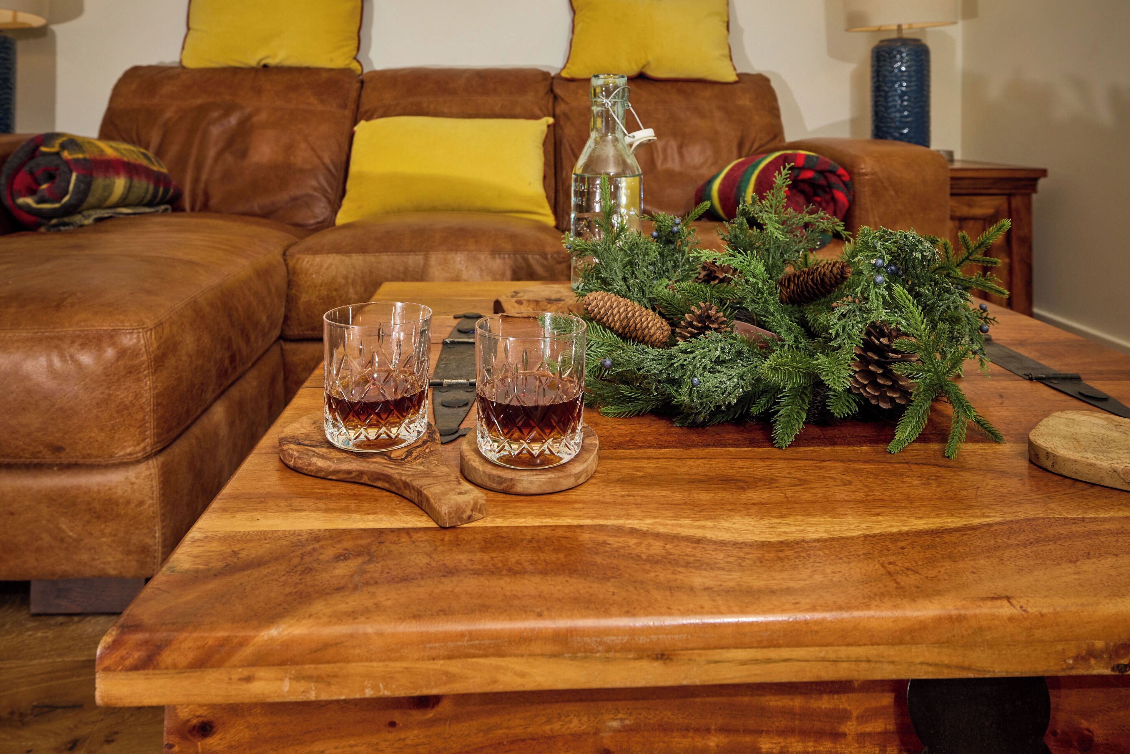A wooden table with two glasses of amber liquid, a bottle, and a pine branch decoration.