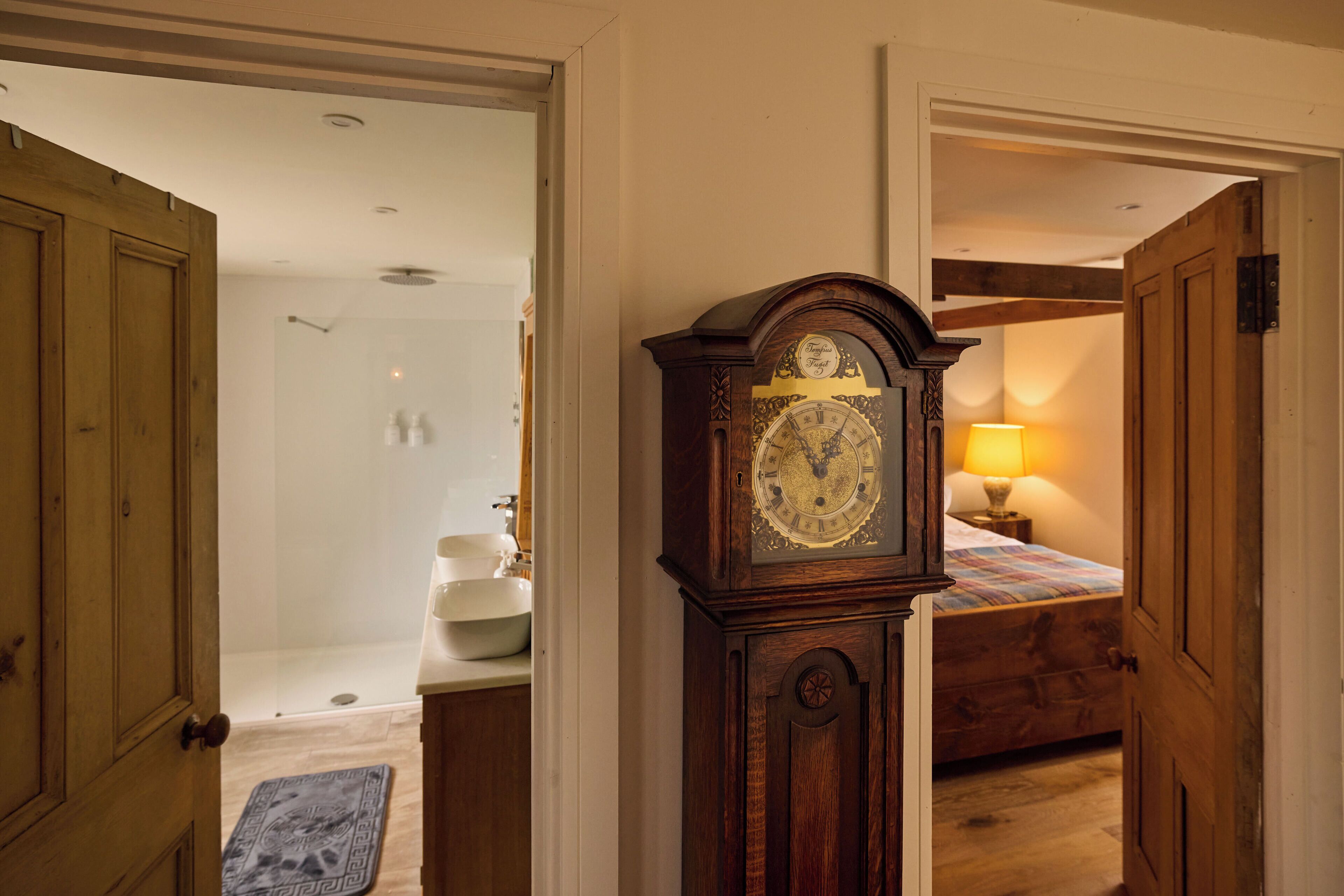 A hallway with a wooden door, a large wall clock, a bathroom with a sink and mirror, and a bed with a lamp.