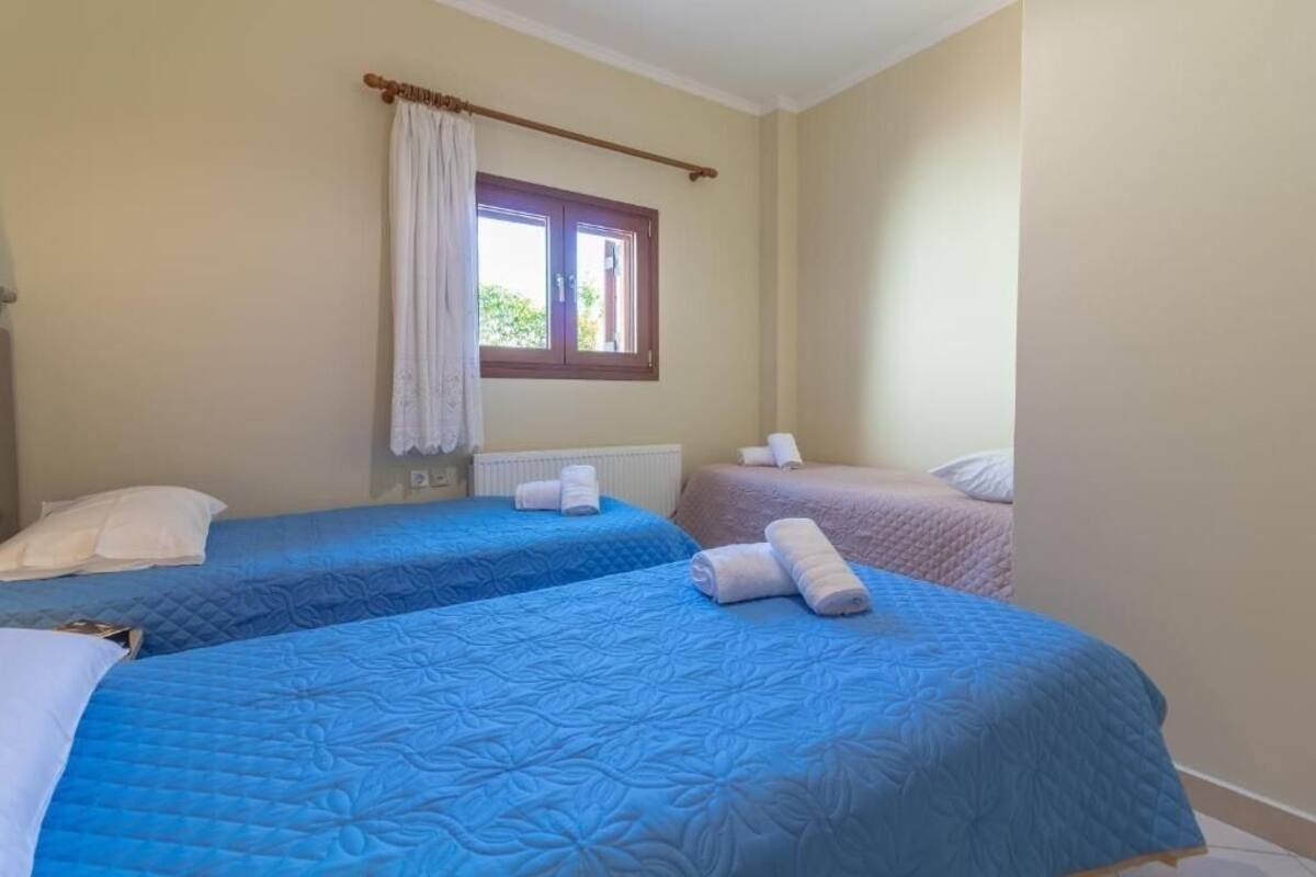 A room with two beds, each with a blue quilted bedspread and white pillows. There is a window with a wooden frame and a white curtain.