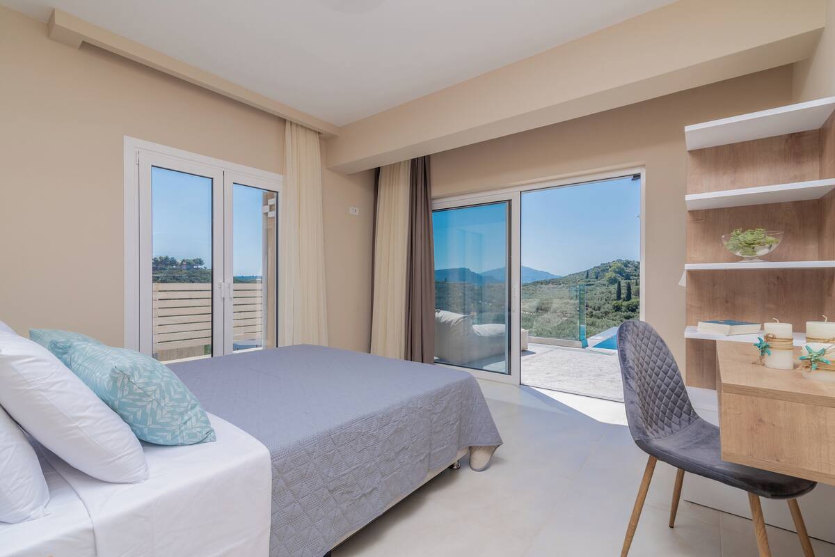 A bedroom with a bed, a chair, a desk, and a balcony with a view of the hills.
