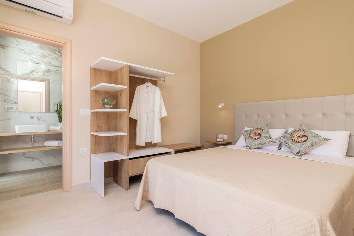 A bedroom with a bed, a bedside table, a nightstand, a wardrobe, and a bathroom with a sink and mirror.