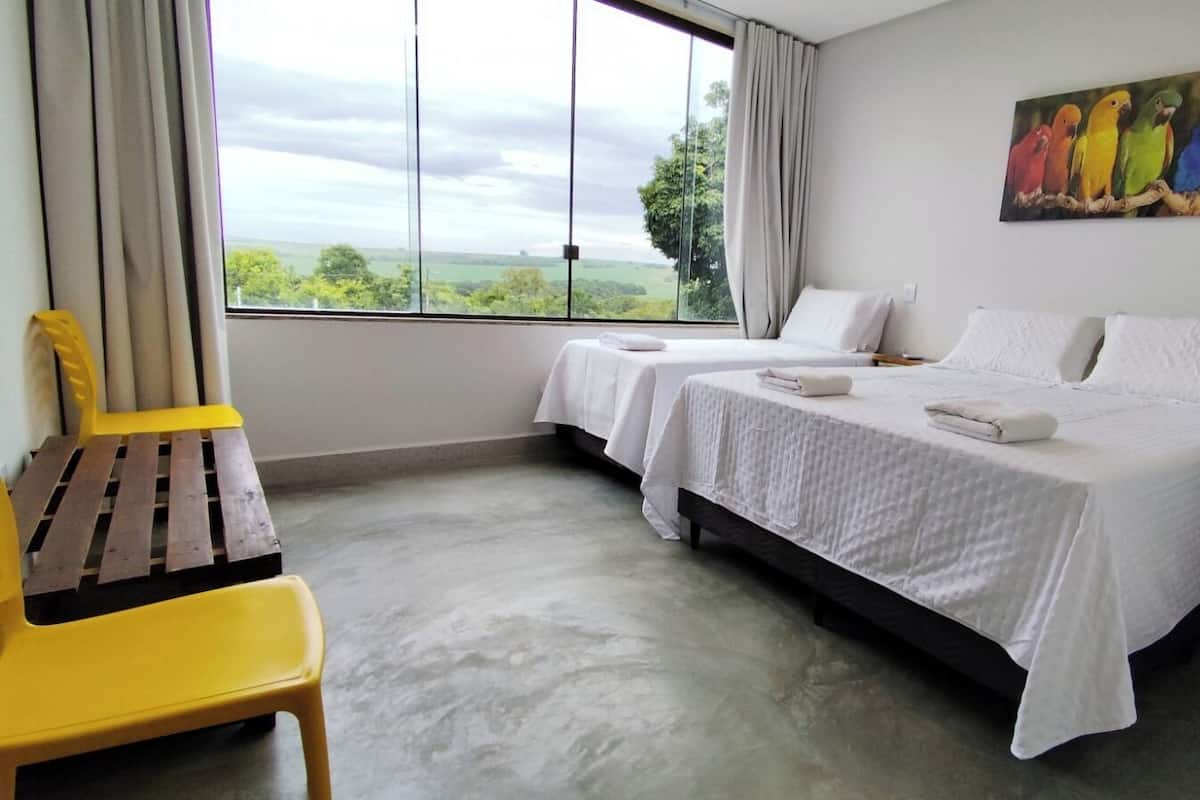A hotel room with two beds, a view of the countryside, a yellow chair, and a painting of colorful birds on the wall.