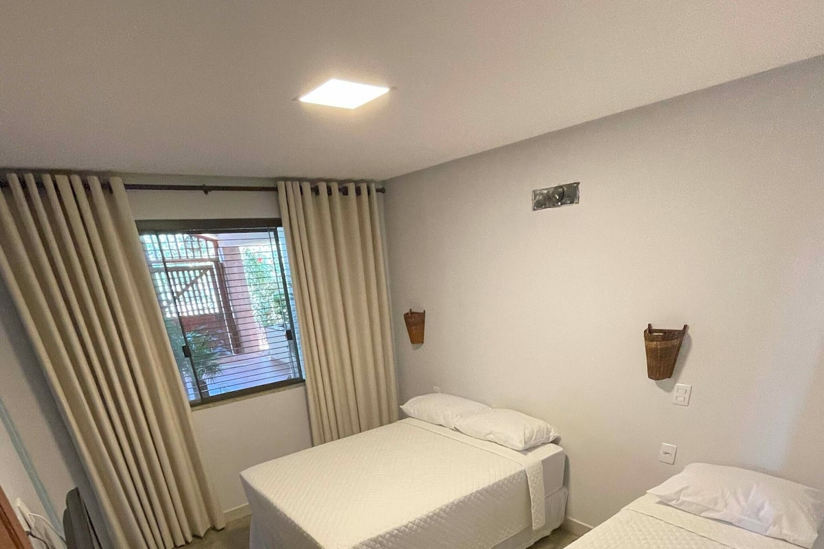 A hotel room with two beds, a window with curtains, a small table, and a wall-mounted light fixture.